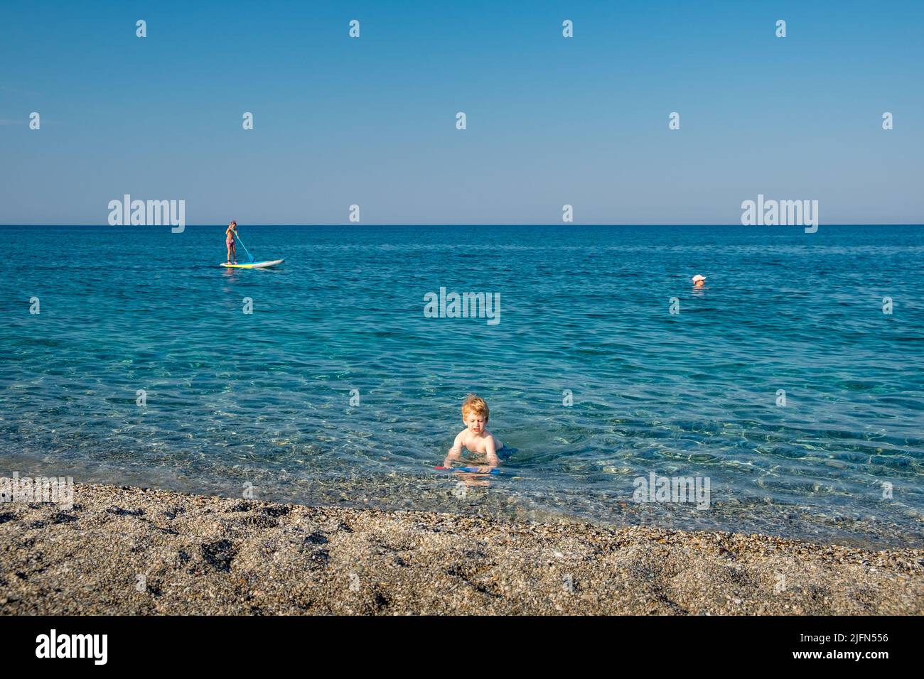 Cute little boy with blond hair playing in the sea with his water gun while other people swimming and stand up paddleboard Stock Photo