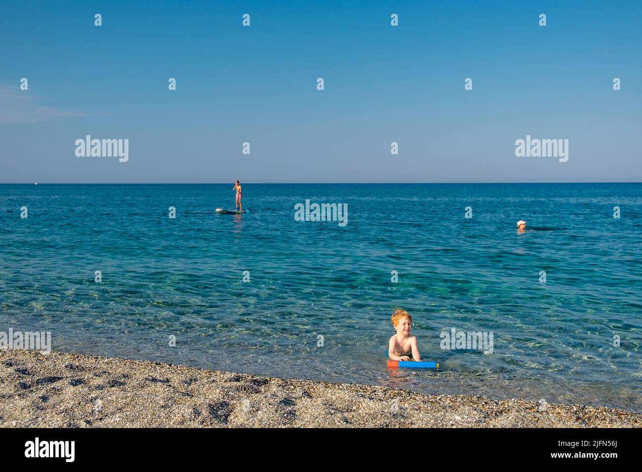 Cute little boy with blond hair playing in the sea with his water gun while other people swimming and stand up paddleboard Stock Photo