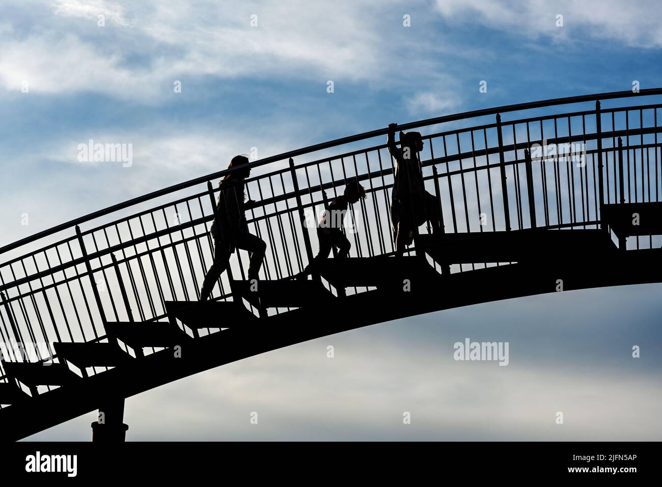 Three children as silhouettes climbing up the steps of a large metal bridge against a blue sky with clouds, future challenge for the next generation c Stock Photo