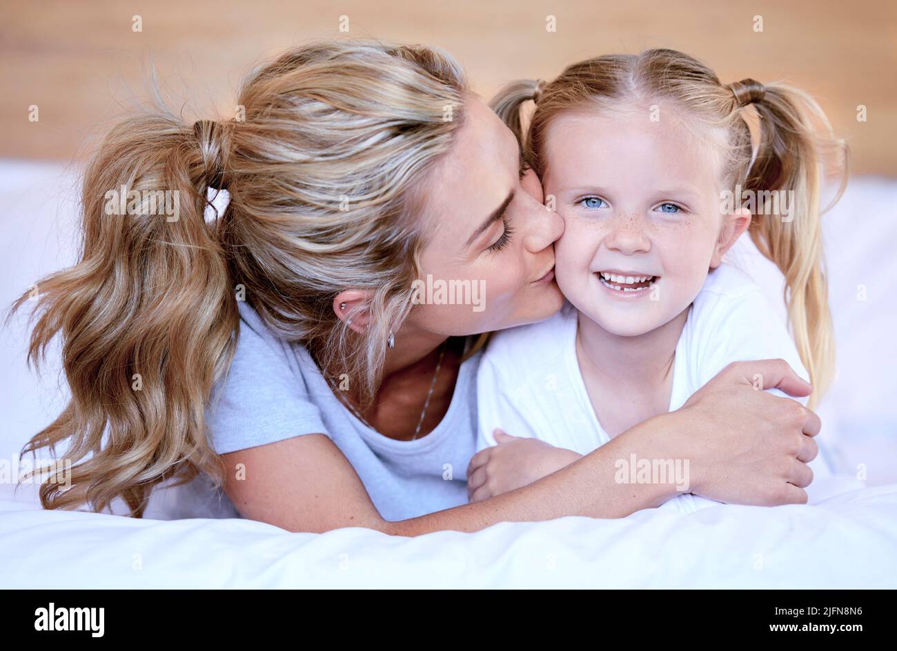 Two blonde caucasian females only looking relaxed and positive while lying in bed together. Young single mother kissing her adorable little daughter Stock Photo