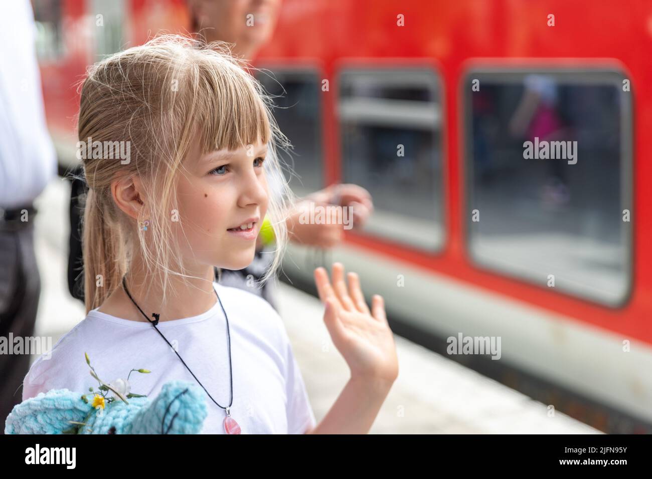 Portait of cute beautiful caucasian little blond kid girl looking waving hand hello goodbye gesture farewell at railway platform station agains red Stock Photo