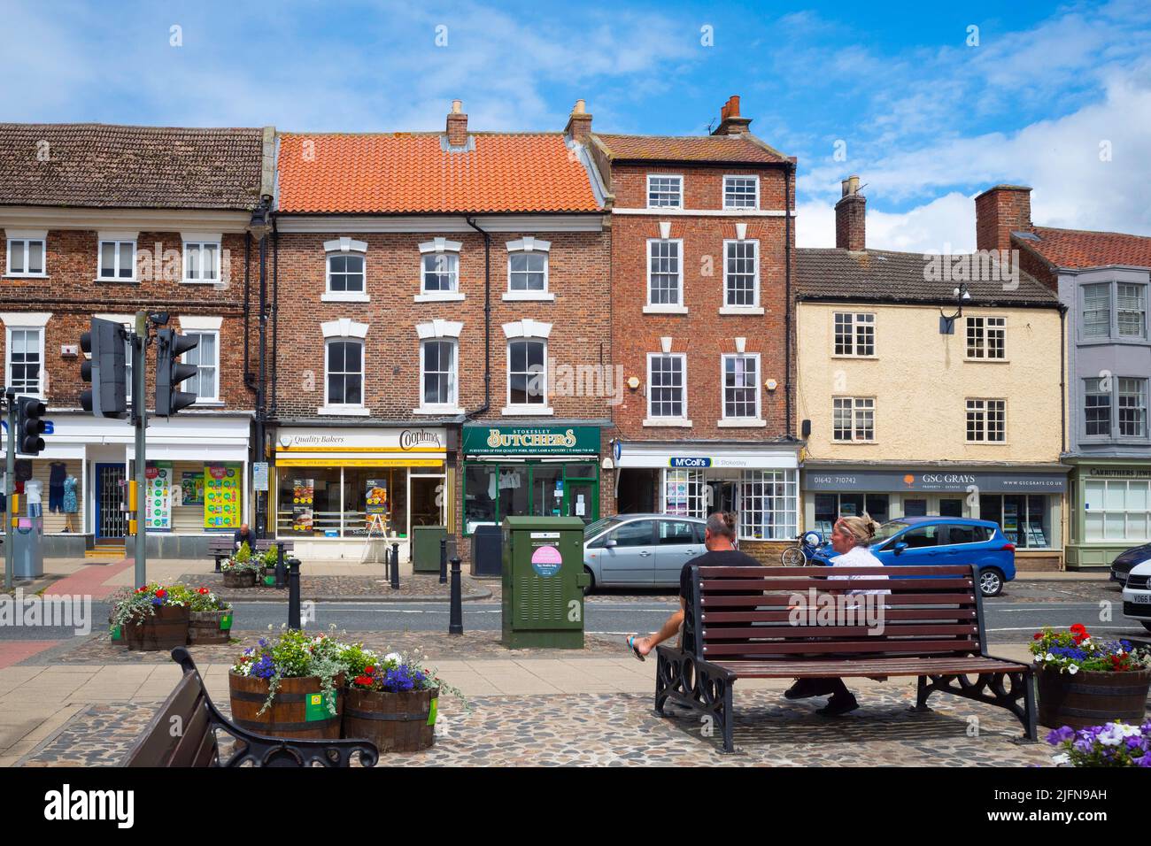 The High Street of the market town of Stokesley North Yorkshire on a sunny summer day with a couple seated on a bench near the road crossing Stock Photo
