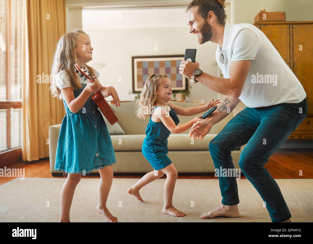 Young happy father singing and dancing with his little daughters in the lounge at home. Cheerful little siblings playing guitar and having fun with Stock Photo