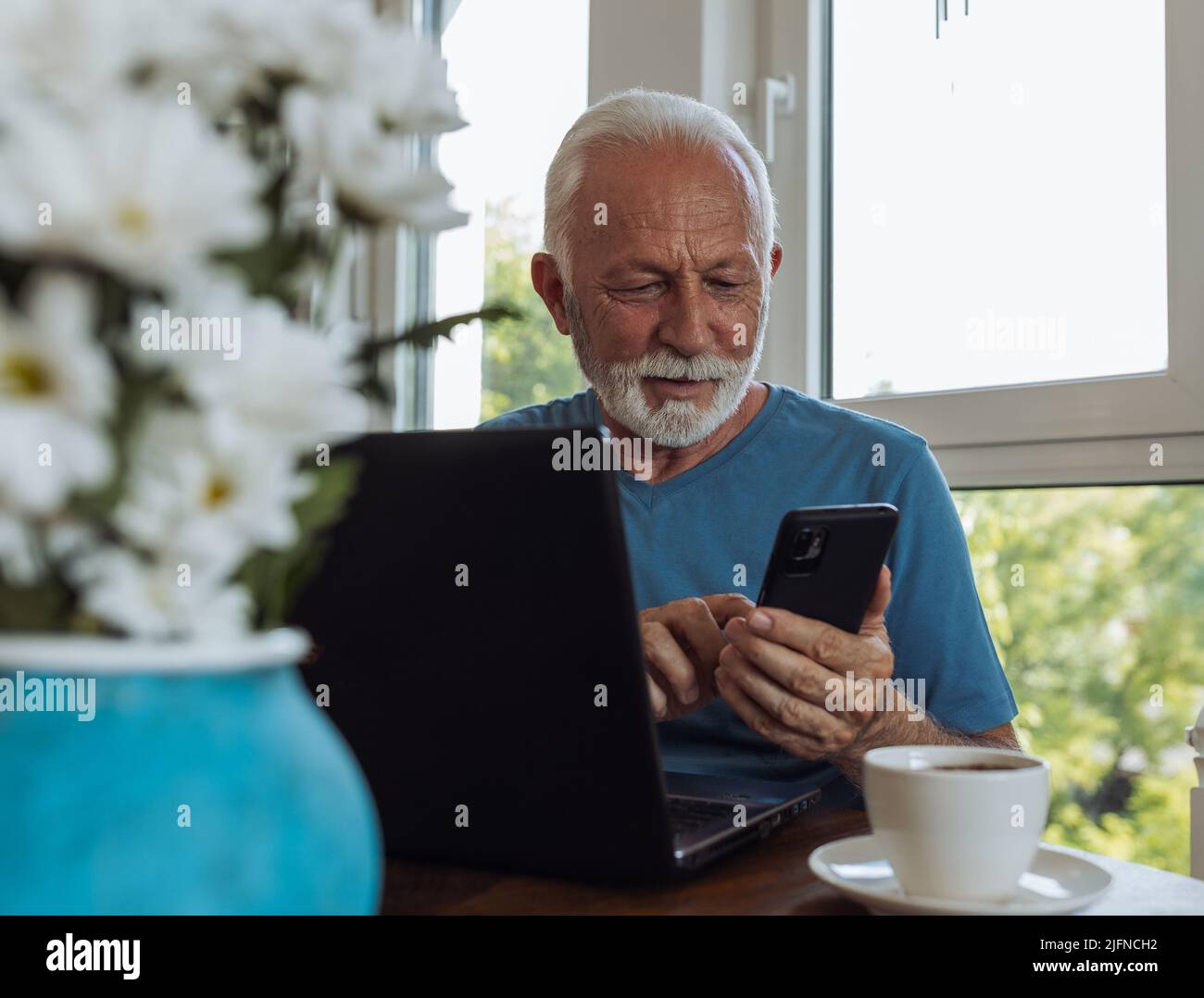 Senior man pensioner working on laptop and smartphone at home at dining table in front of window, online shopping with mobile application Stock Photo