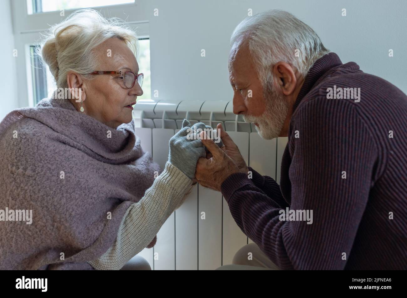 Senior couple holding each other hands beside heater in living room and trying to warm in winter season and energy crisis Stock Photo