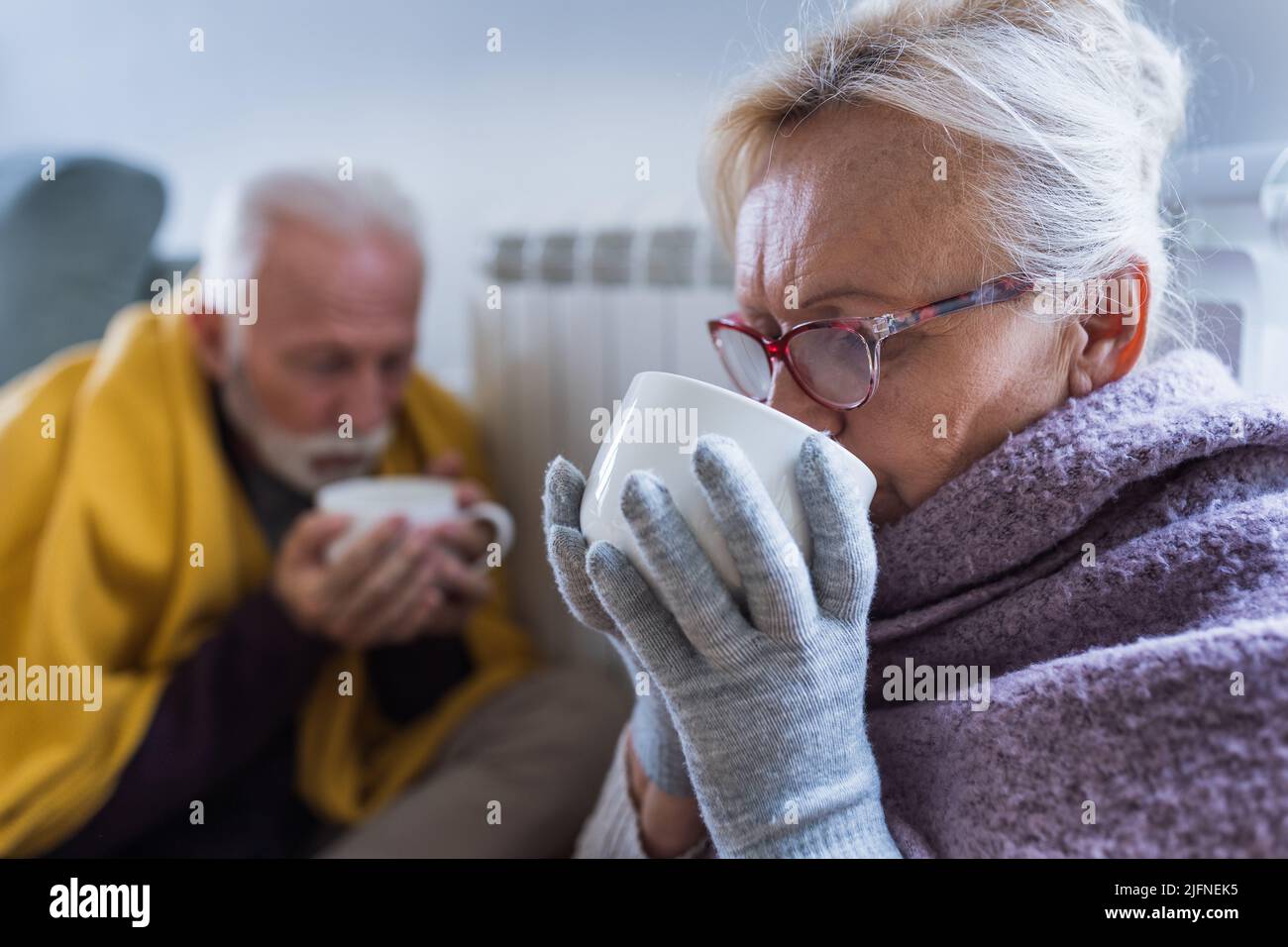 Senior couple covered with blanket sitting beside heater in living room, drinking hot tea from big cup and trying to warm in winter season and energy Stock Photo