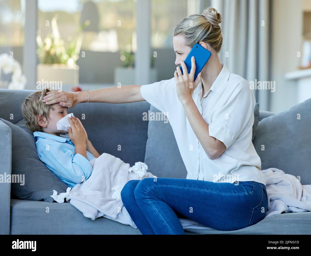 Young caucasian mother feeling her sick sons forehead while on a call using a phone sitting on the couch in the lounge at home. Ill child blowing his Stock Photo