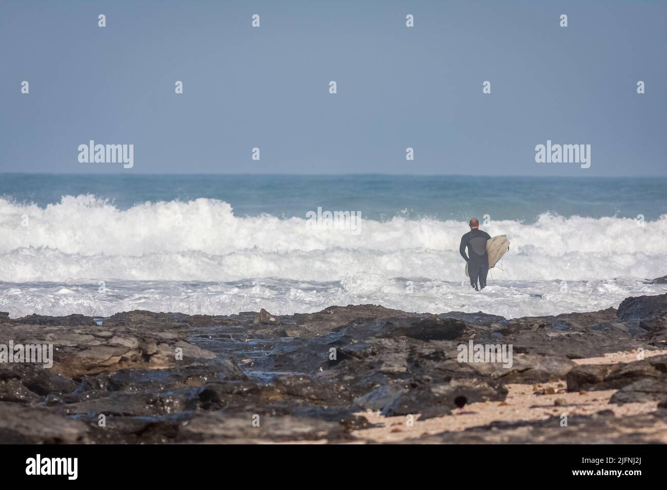 A back view of a surfer in a wetsuit with surfboard walking into the