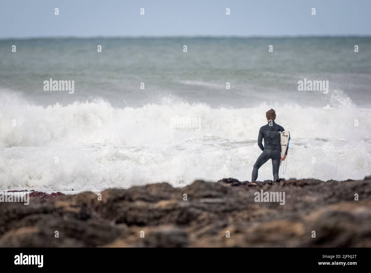A back view of a surfer in a wetsuit with surfboard standing by the