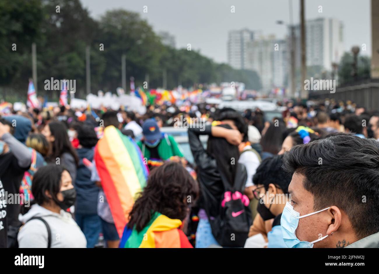 Selective focus photo of a young Latinx man with a mask gazing out to the street full of people and flags at Lima's Marcha del Orgullo, pride march. Stock Photo