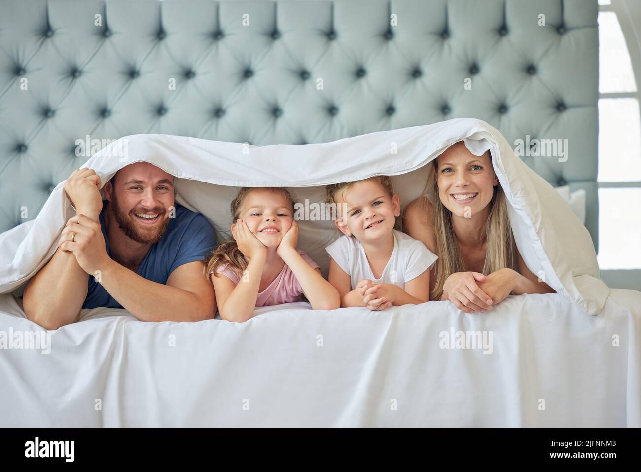 Portrait of happy parents with little children lying on bed at home under blanket. Caucasian girls bonding with their mother and father. Smiling young Stock Photo