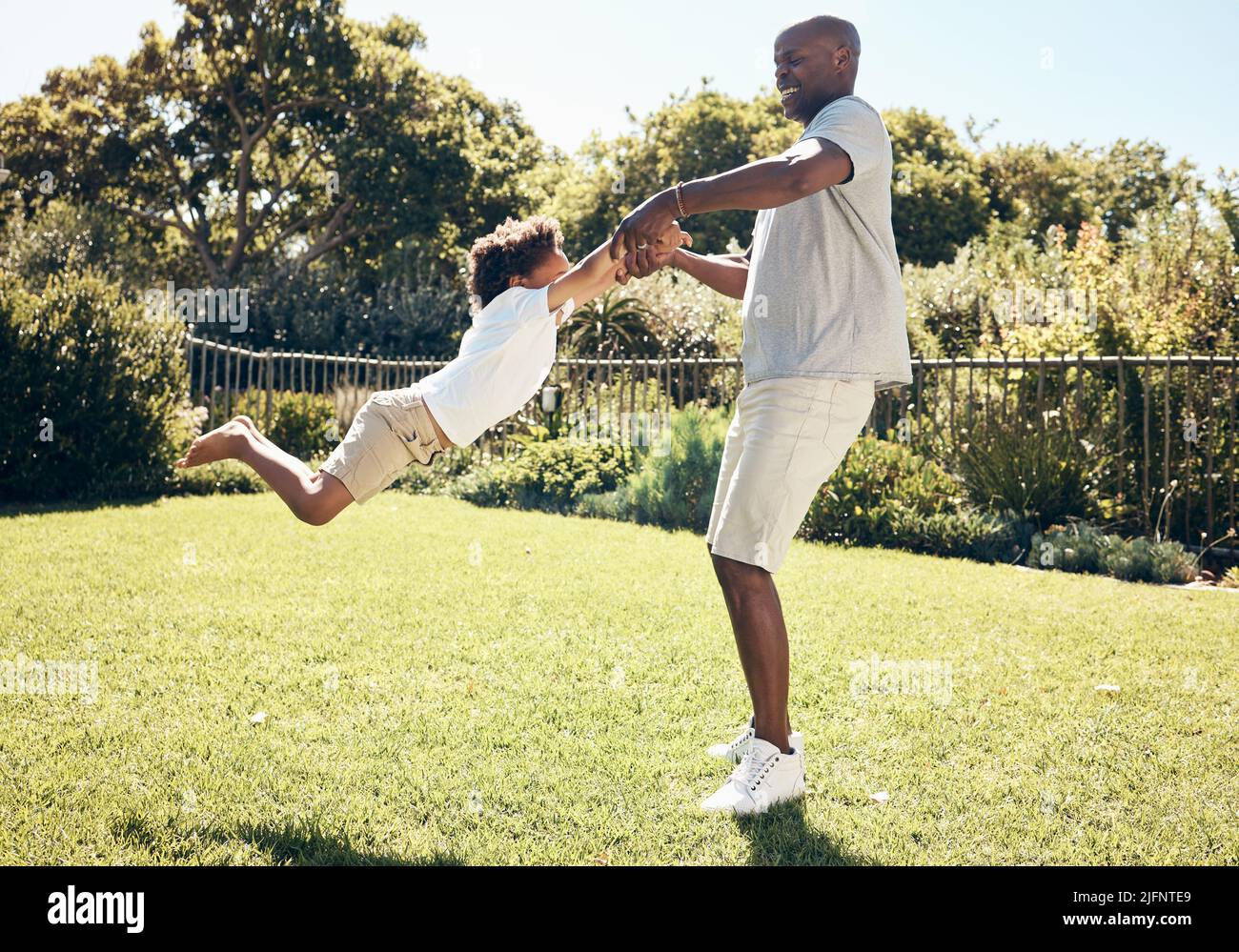 Cheerful african american dad swinging his son by the arms. Father playing with his son while spending time together outdoors at the park or in their Stock Photo