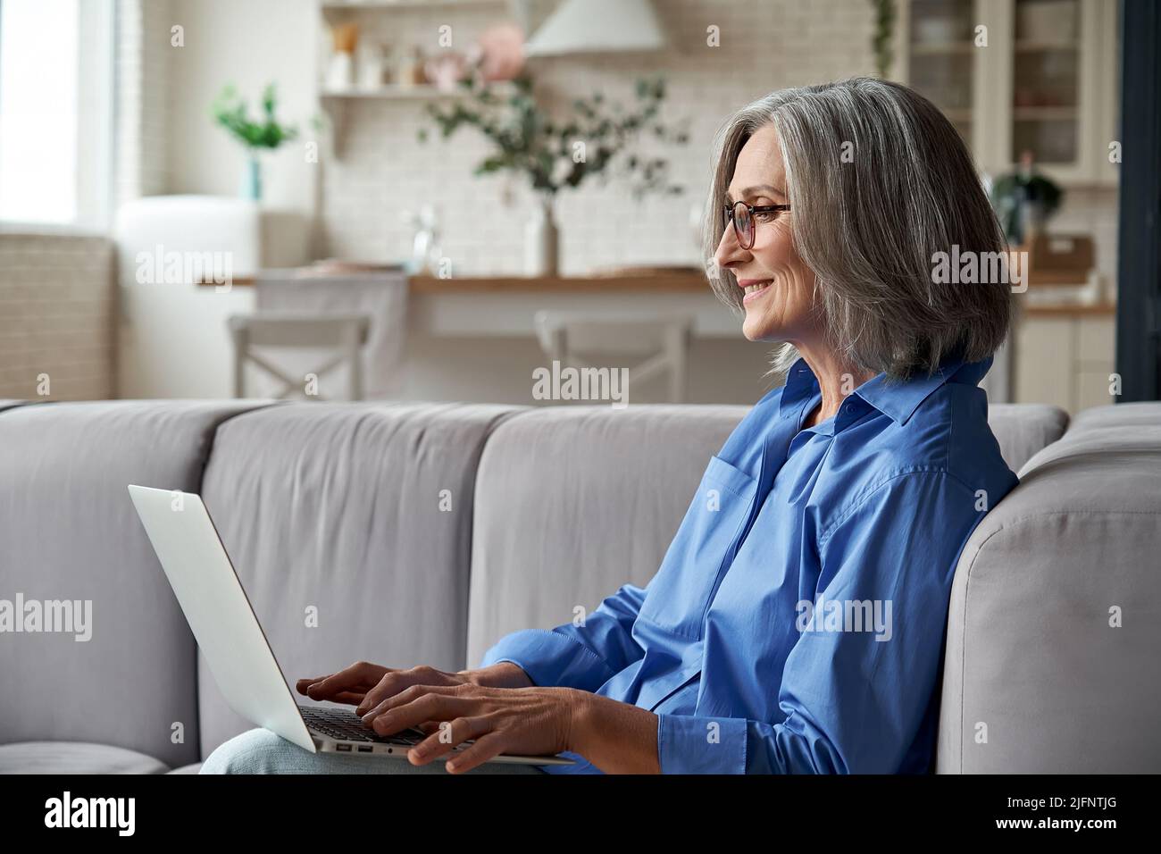Happy 60s mature woman using laptop computer sitting on couch at home. Stock Photo