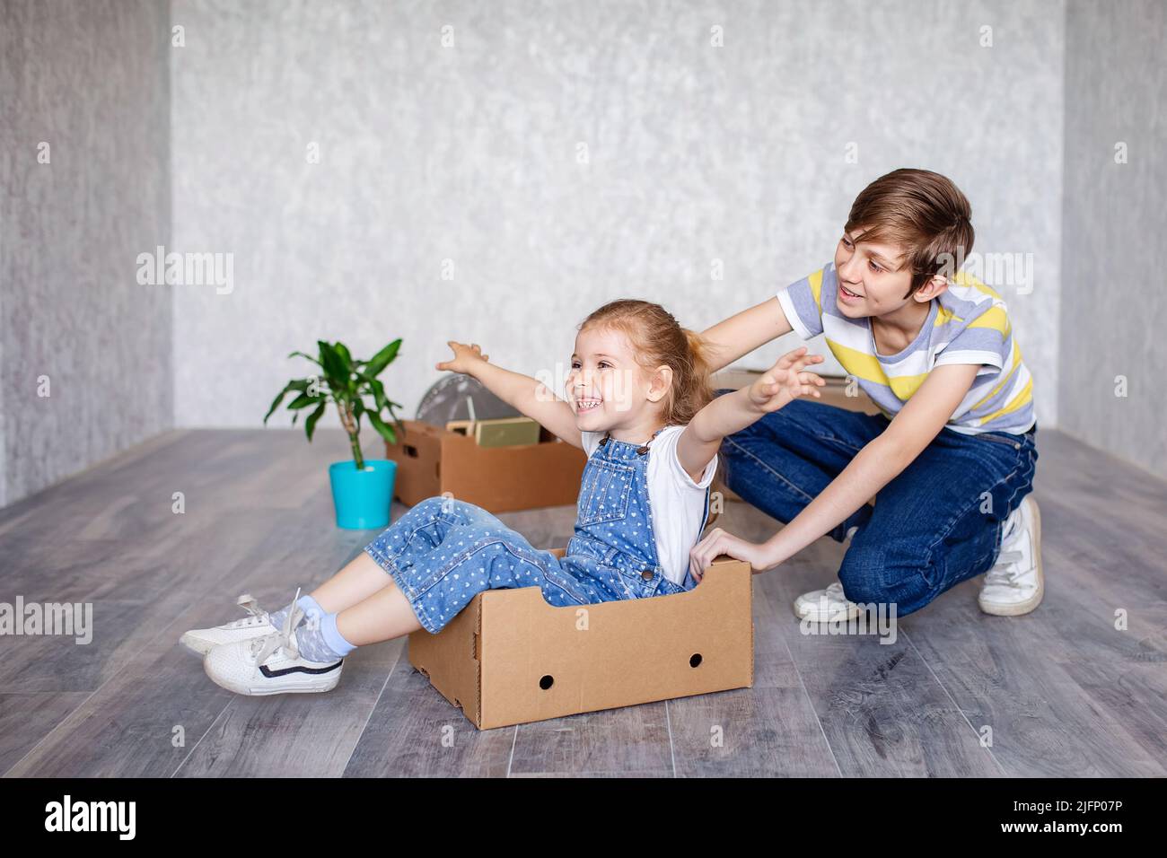 Brother rides sister in a cardboard box. Children play with boxes at home in quarantine. A boy and a girl have moved into a new apartment and are Stock Photo