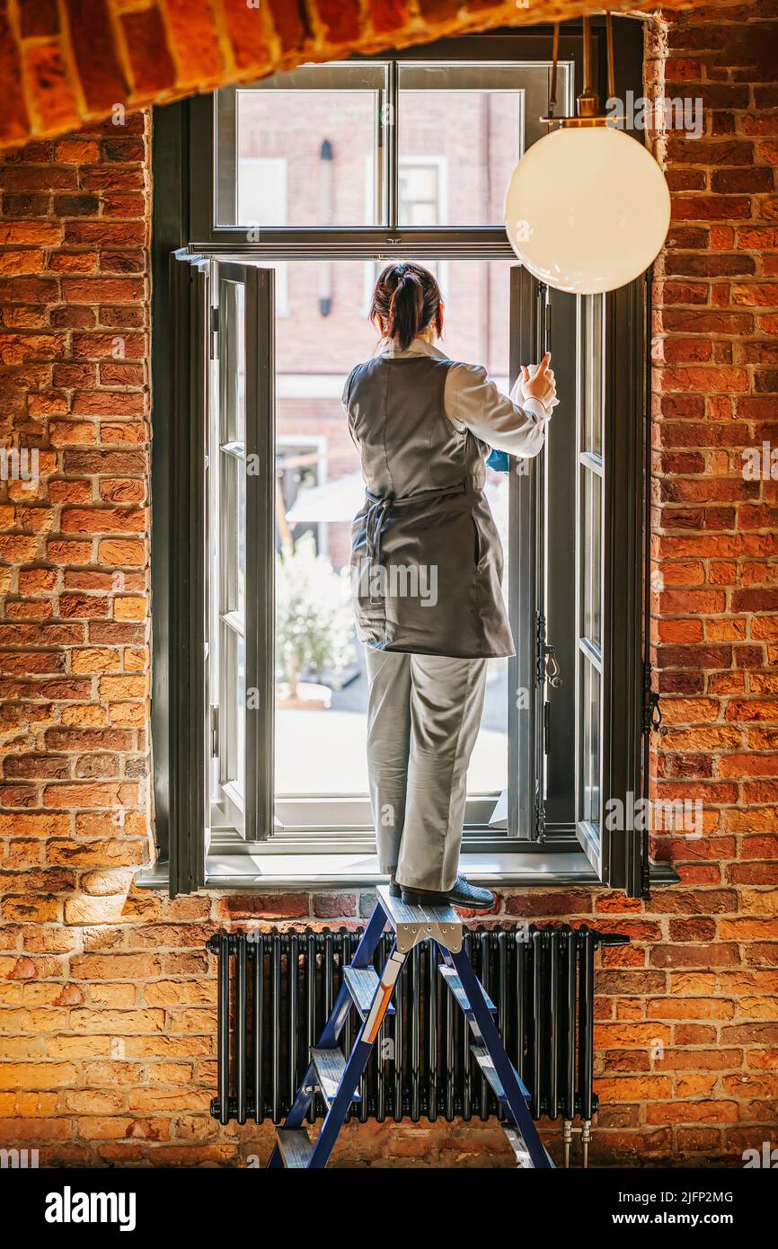 Woman worker washes window while standing with her back to us on stepladder Stock Photo