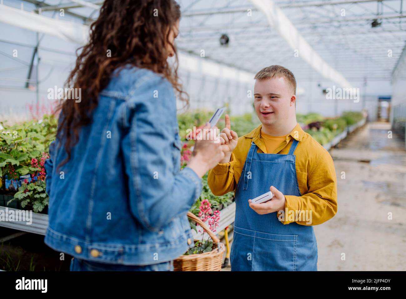 Happy young employee with Down syndrome working in garden centre, taking payment from customer. Stock Photo