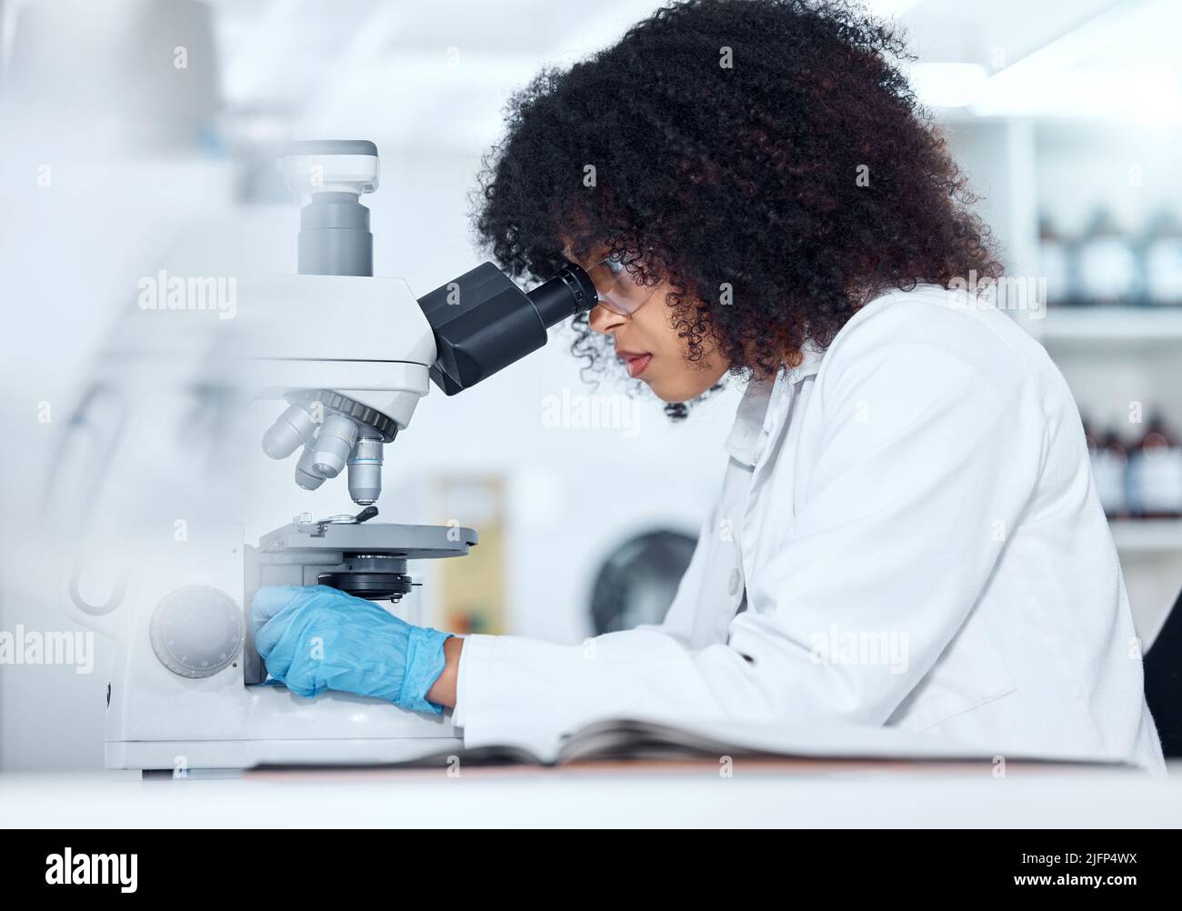 One mixed race scientist with curly hair wearing safety goggles and gloves analysing medical test samples on a microscope in a lab. Young woman doing Stock Photo