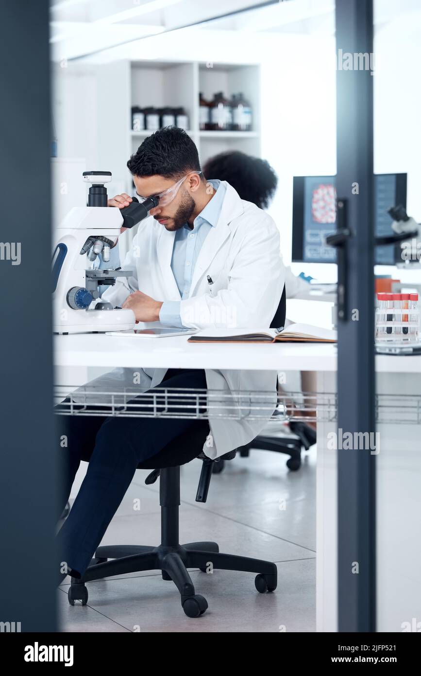 One mixed race scientist wearing safety goggles while analysing medical test samples with a microscope in a lab. Young indian man investigating with Stock Photo