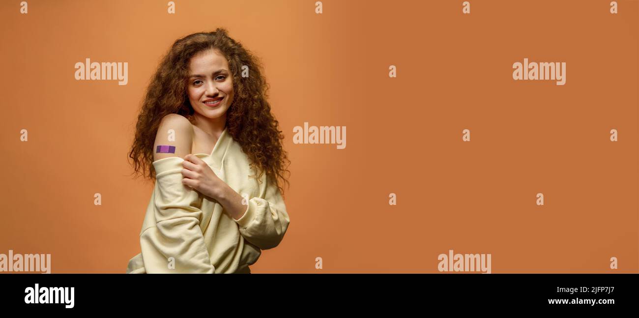Portrait of a female smiling after getting a vaccine. Woman holding down her shirt sleeve and showing her arm with bandage after receiving vaccination Stock Photo