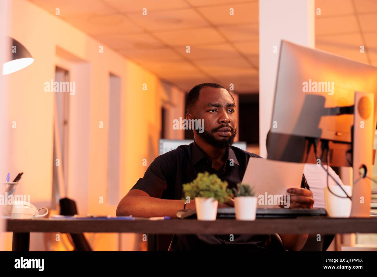 Executive manager analyzing paperwork report and taking notes to create research presentation in office with big windows. Working on project data with documents and computer during sunset. Stock Photo