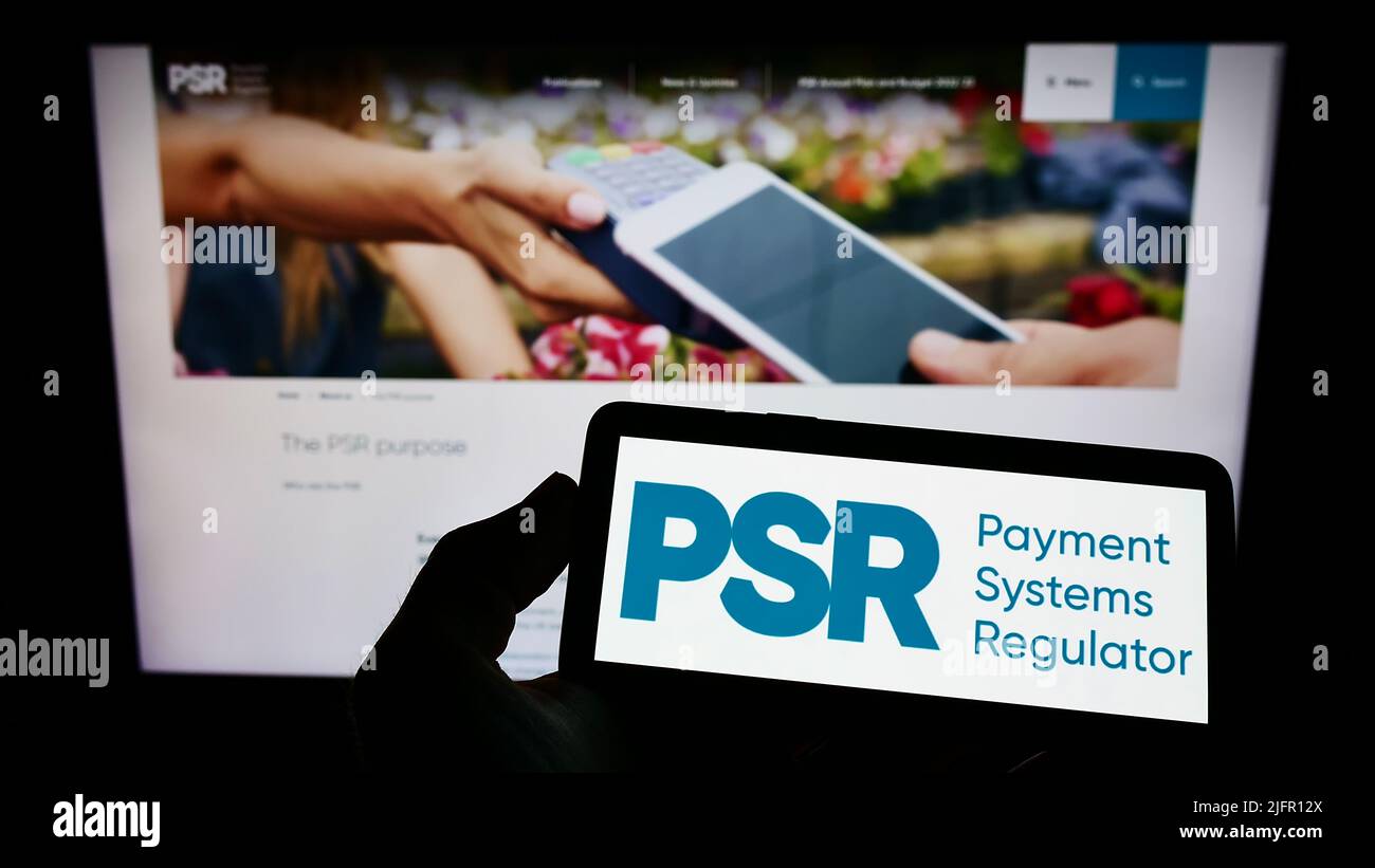 Person holding smartphone with logo of British Payment Systems Regulator (PSR) on screen in front of website. Focus on phone display. Stock Photo