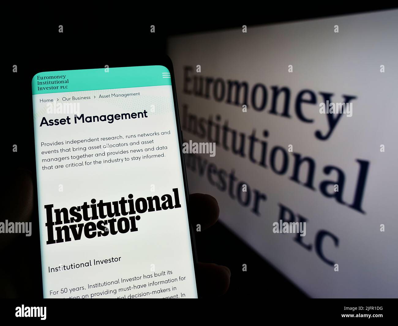 Person holding cellphone with webpage of company Euromoney Institutional Investor plc on screen with logo. Focus on center of phone display. Stock Photo