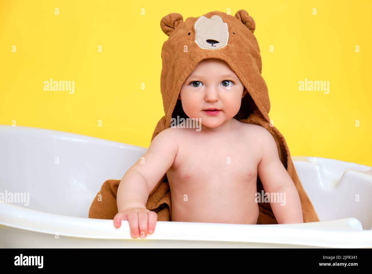 Happy baby toddler boy with hood bathrobe in a white bathtub on a studio yellow background. A
