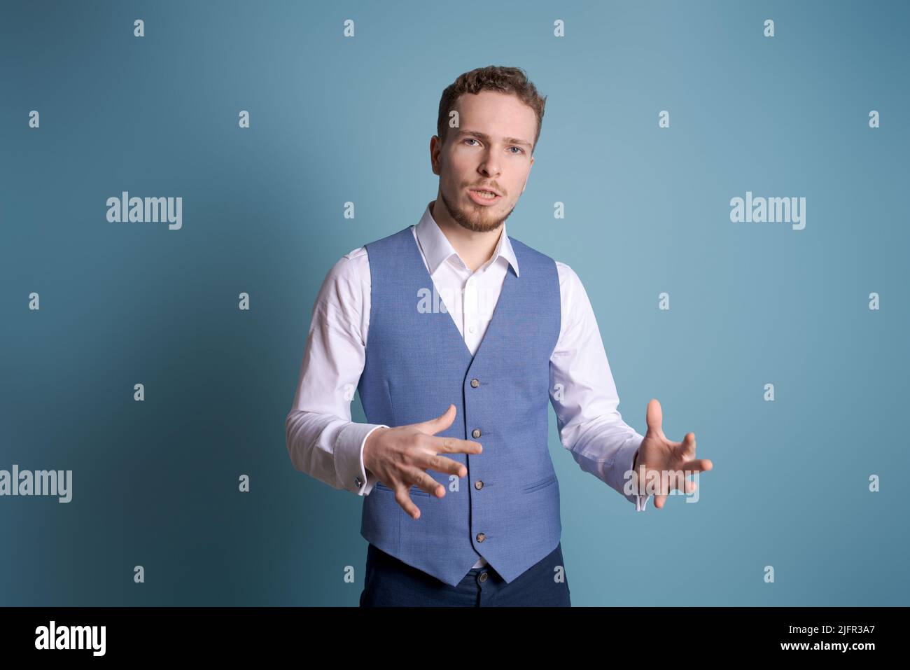 Young handsome man with beard in white shirt and blue vest standing on blue background is confused and wonders. Unclear with doubt, thinking gesticulating with hands. Thoughtful concept. Stock Photo