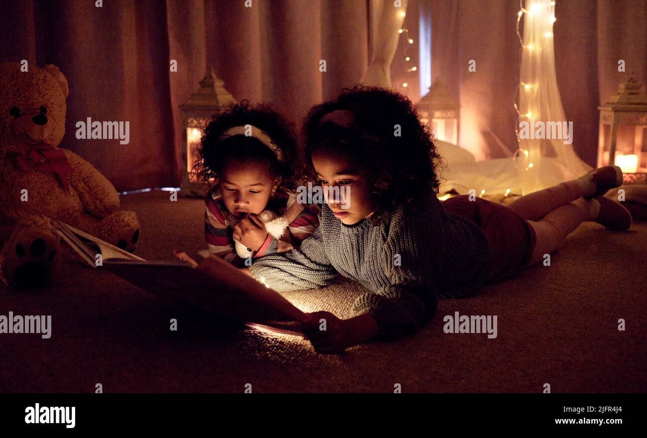 Theres so much magic in a book. Shot of two adorable little girls reading a book together in a room at night. Stock Photo