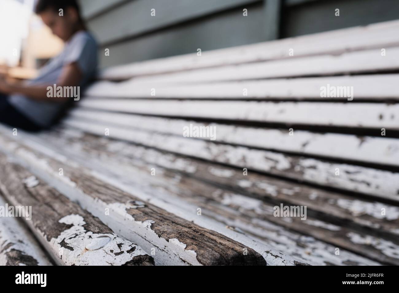 Abandoned bench, details of peeling white paint and wooden material.Silhouette of a child sitting with his head bowed.Concept for bullying, loneliness Stock Photo