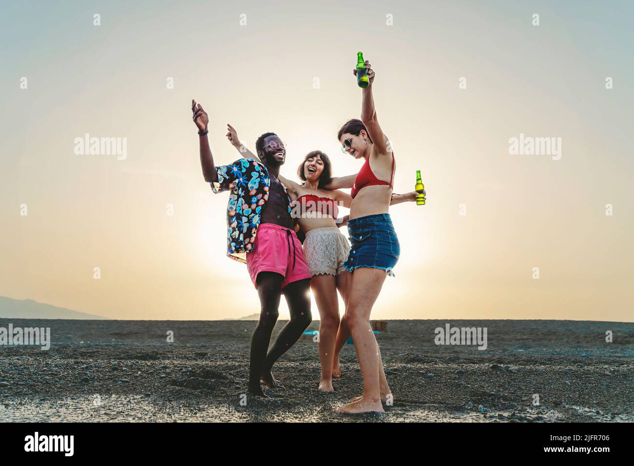 Happy multiracial group of young people dancing crazy on the beach at sunset drinking bottled beers - multiethnic people, fun, friendship alcohol and Stock Photo