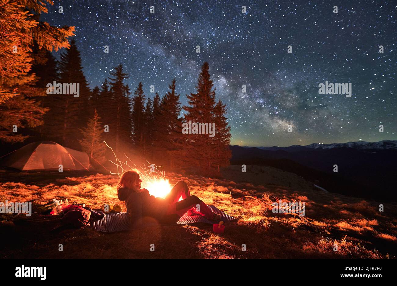 Female hiker lying at karemat near bonfire admire beauty of stunning starry sky during night in campsite, backpacker visiting mountains with tent near spruce forest in National Park Stock Photo