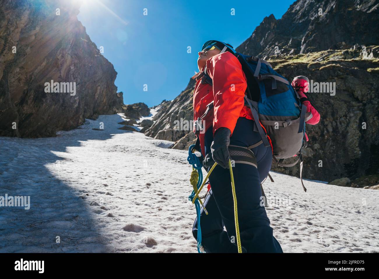 Team roping up woman dressed high altitude mountaineering clothes and harness climbing with backpack by snowy slope in the couloir with backlight sun Stock Photo