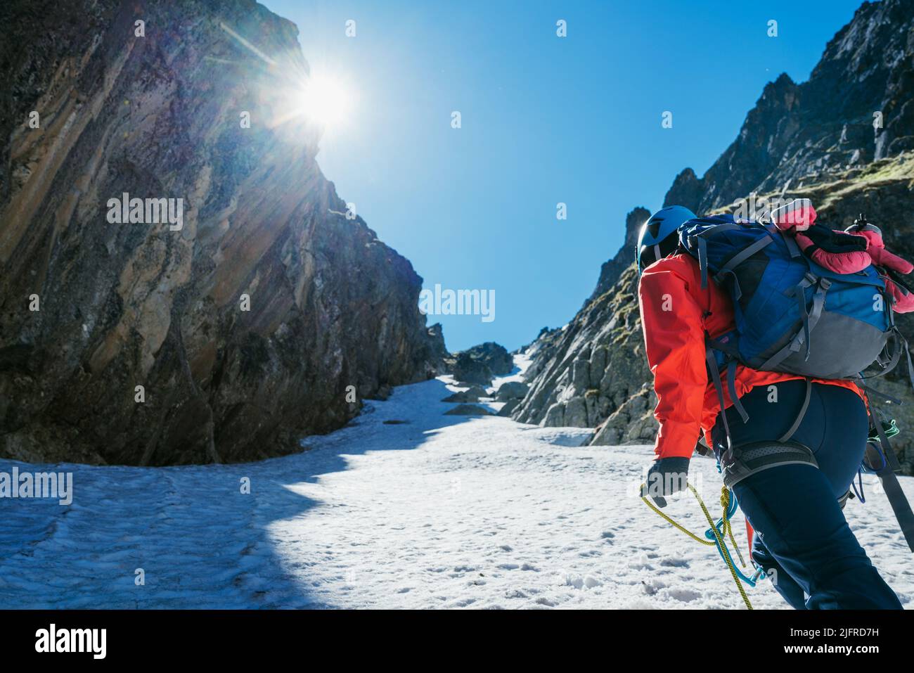 Team roping up woman dressed high altitude mountaineering clothes and harness climbing with backpack by snowy slope in the couloir with backlight sun Stock Photo