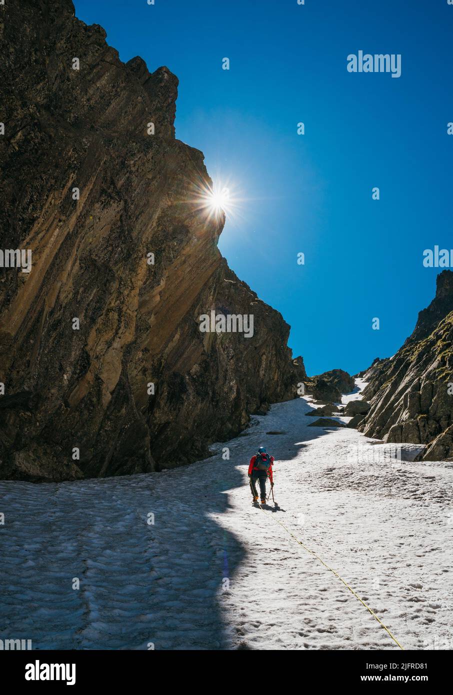 Team roping up woman with climbing axe dressed high altitude mountaineering clothes with backpack walking by snowy slope in the couloir with backlight Stock Photo