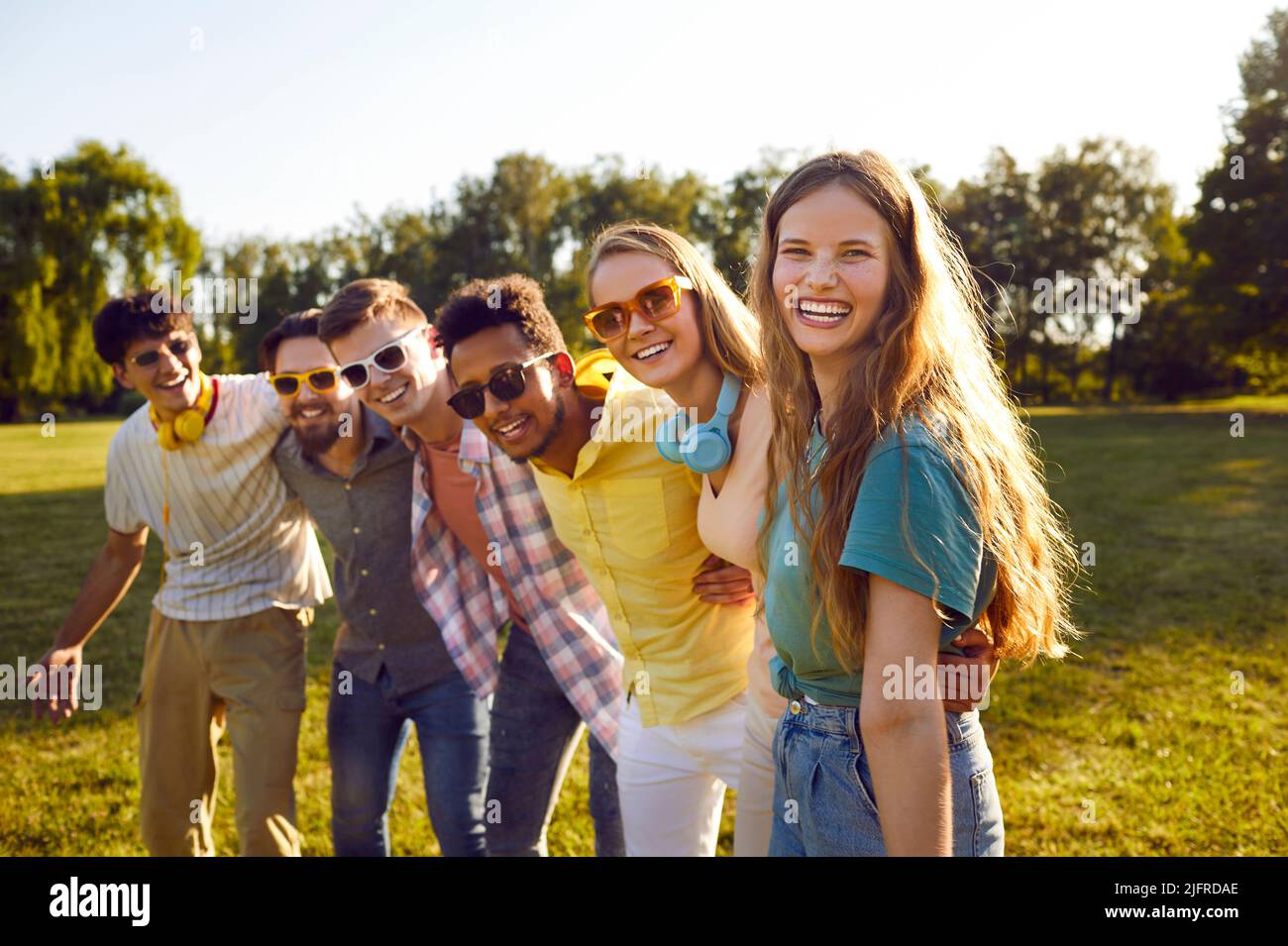 Portrait of happy excited young woman standing with her multiracial friends in green park. Stock Photo