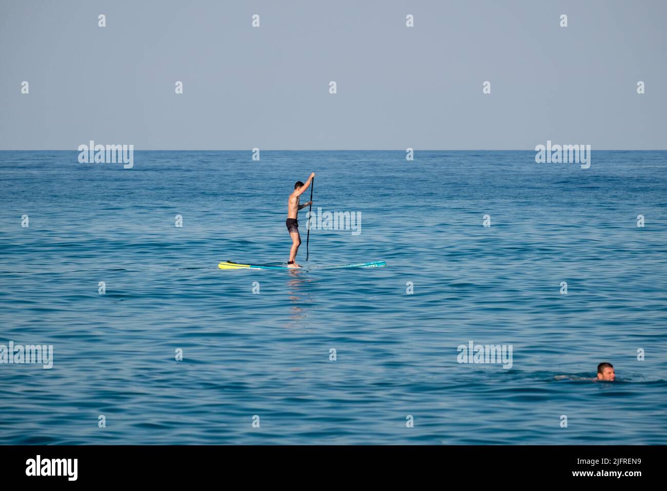 A young man on stand up paddleboard and another man swimming in the calm waters of Aegean sea Stock Photo