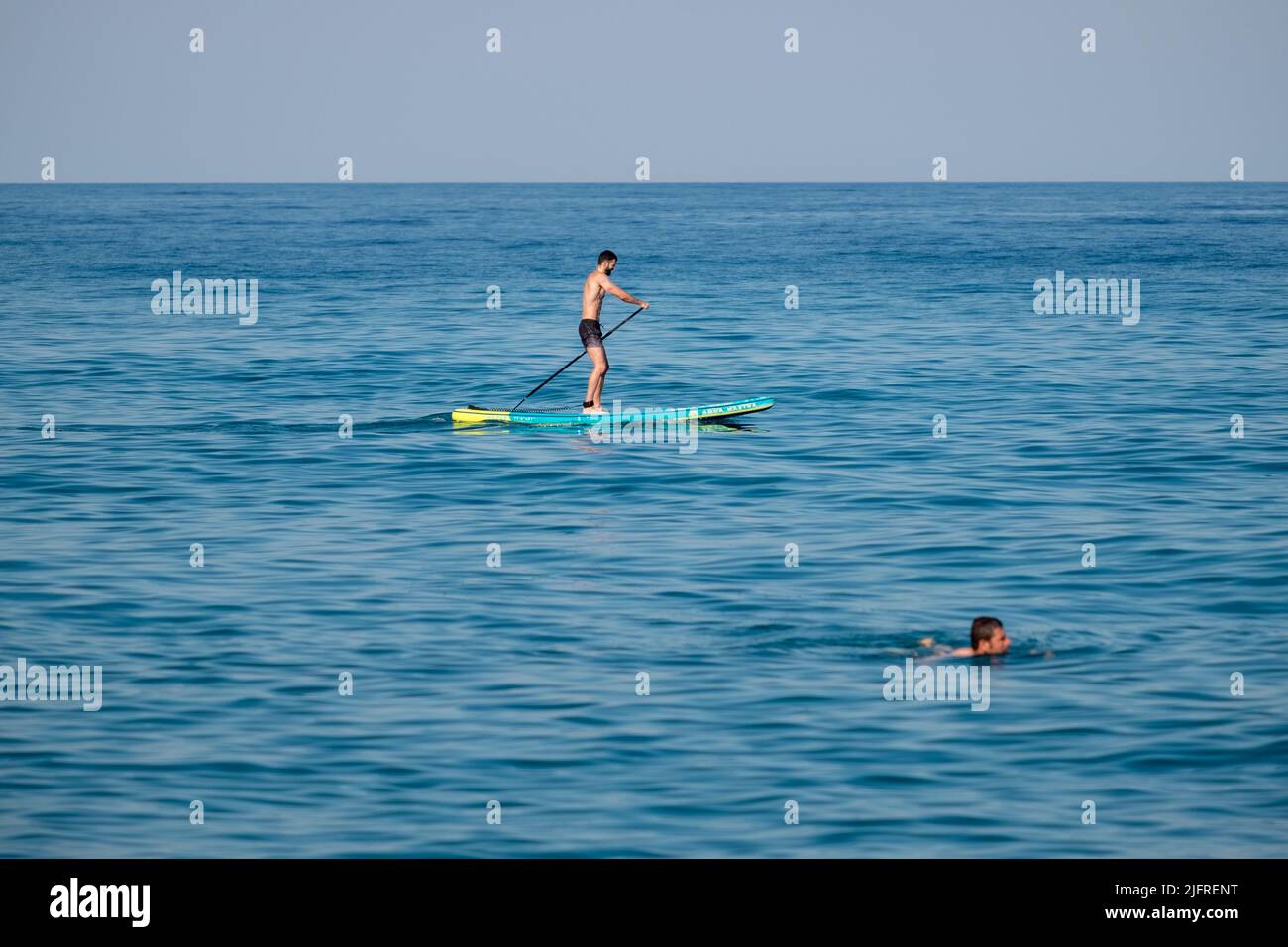 A young man on stand up paddleboard and another man swimming in the calm waters of Aegean sea Stock Photo
