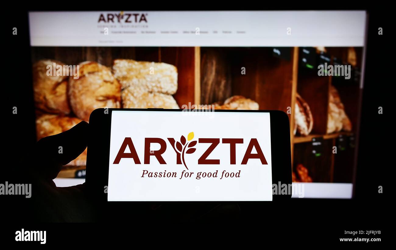 Person holding smartphone with logo of Swiss food company Aryzta AG on screen in front of website. Focus on phone display. Stock Photo