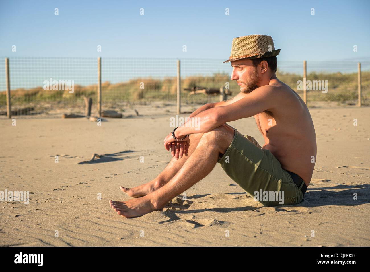 Young boy looking out to sea - Young man wearing sun hat and sitting outdoors watching the sunset. Concept of thinking and relaxation. Stock Photo