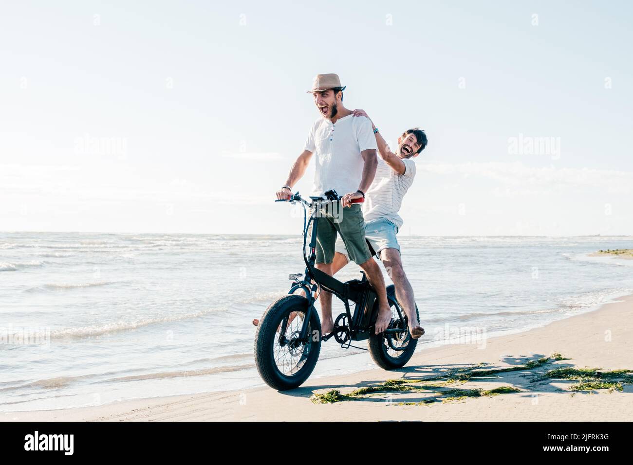 Cheerful friends couple on bicycle having fun with electric bike - carefree boys having fun and smiling on bicycle on the beach on a sunny day Stock Photo