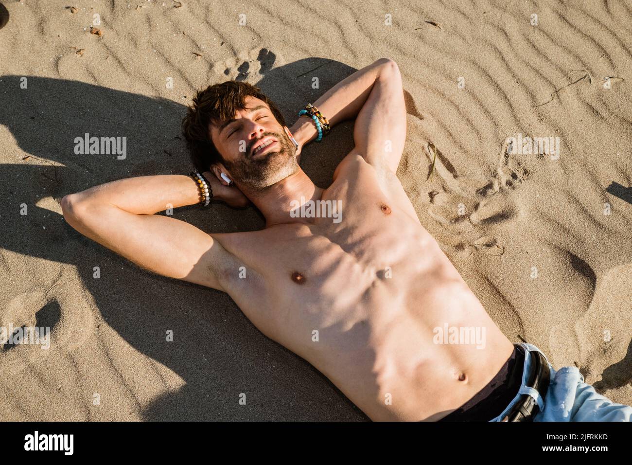 Young boy listening favorite music with earphones on beach - Happy attractive man lying down on sand listening to music on a smartphone Stock Photo