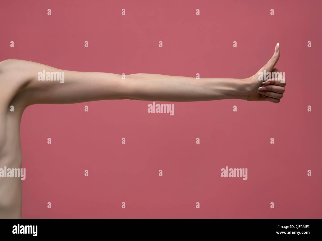 Young woman's stretched skinny arm thumb up. Isolated on pink