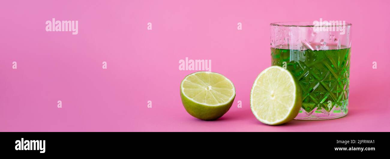 green alcohol drink in faceted glass with water drops near limes on pink, banner Stock Photo