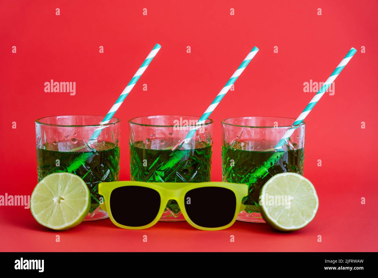 green alcohol drink in faceted glasses with straws and halves of limes near sunglasses on red Stock Photo