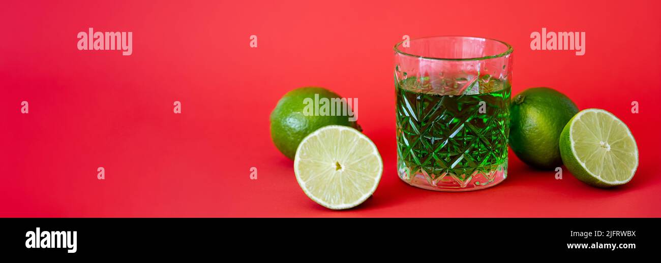 green alcohol drink in faceted glass near sour limes on red, banner Stock Photo