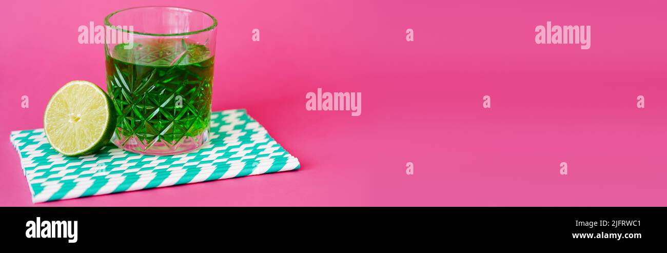 glass of green alcohol drink on striped paper straws near sliced lime on pink, banner Stock Photo