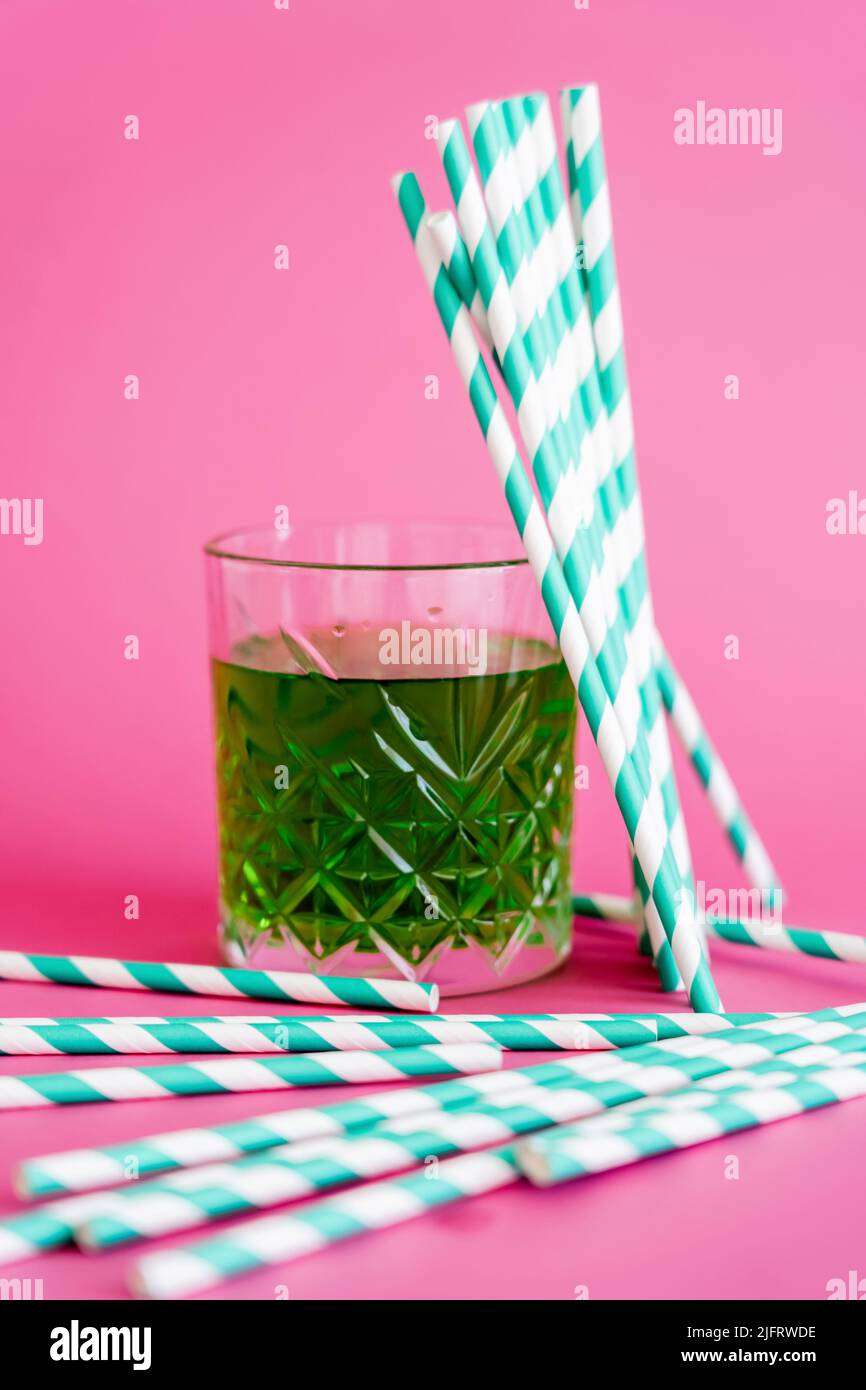 faceted glass of green alcohol drink near striped paper straws on pink Stock Photo