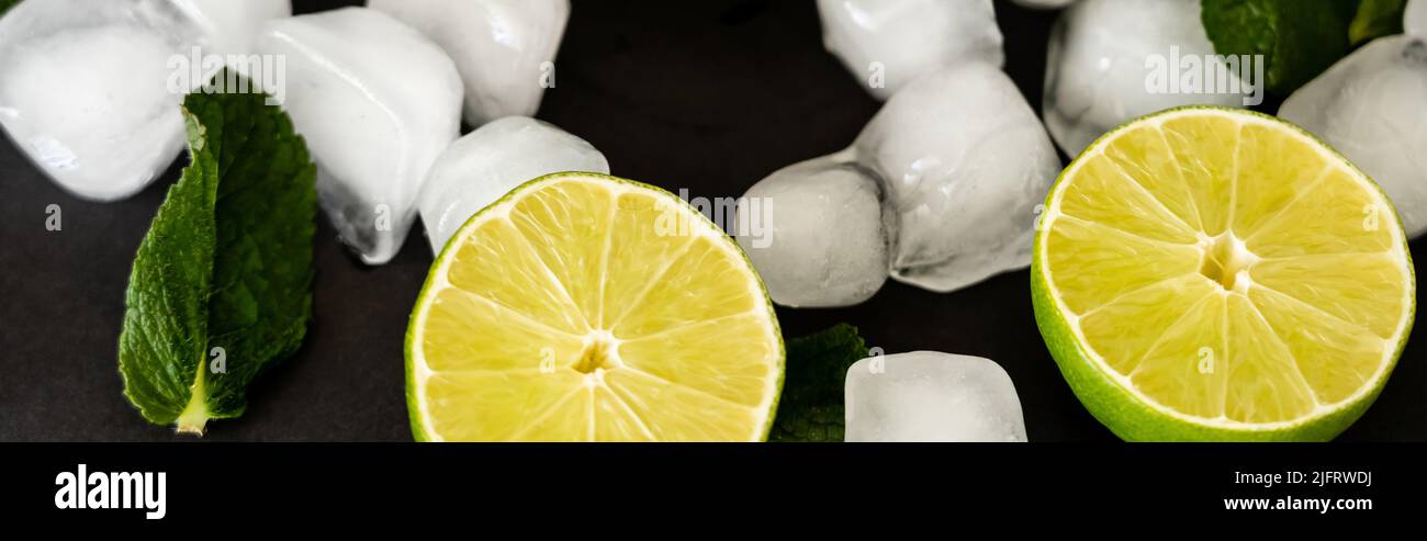 high angle view of melting ice cubes near sliced limes and milt leaf on black background, banner Stock Photo