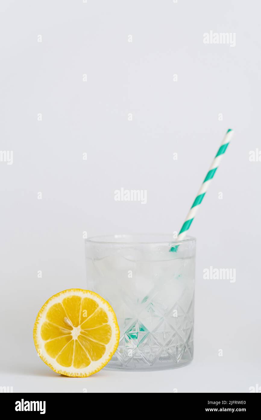 faceted cold glass with ice cubes and paper straw near sliced lemon on white Stock Photo